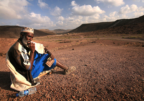 1st Place Photojournalism by Sgt. Alex Kleinsmith: A nomadic Djiboutian snacks on crackers while watching Marines from Battery A, 3rd Low Altitude Air Defense Battalion during a live-fire training exercise in the mountains of Djibouti, Africa. Spent shell casings made of brass are valued by the Djiboutian populace, who trade in the shells for a small, albeit vital payoff.